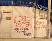 Image of a white cement wall with barbed wire on top of it, covered with red and black graffiti in Spanish that says: “The crime is 1954. No one is illegal on stolen land. Marco wants to be Rubio (blonde) just like Trump.”