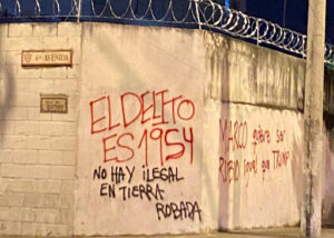Image of a white cement wall with barbed wire on top of it, covered with red and black graffiti in Spanish that says: “The crime is 1954. No one is illegal on stolen land. Marco wants to be Rubio (blonde) just like Trump.”