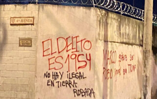 Image of a white cement wall with barbed wire on top of it, covered with red and black graffiti in Spanish that says: “The crime is 1954. No one is illegal on stolen land. Marco wants to be Rubio (blonde) just like Trump.”