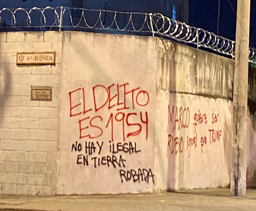 Image of a white cement wall with barbed wire on top of it, covered with red and black graffiti in Spanish that says: “The crime is 1954. No one is illegal on stolen land. Marco wants to be Rubio (blonde) just like Trump.”