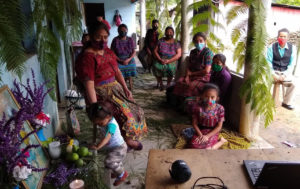 In the front of a house, 9 people, mostly women, all using facial masks are sitting in the distance while they watch the livestream of the assembly on a laptop. There is a small altar on the left side, where a child plays.