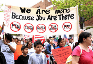 imagen de un grupo de personas manifestando. La multitud se compone principalmente de mujeres y niñxs. Se sostiene una pancarta con las palabras "Estamos aquí porque tú estás allí".