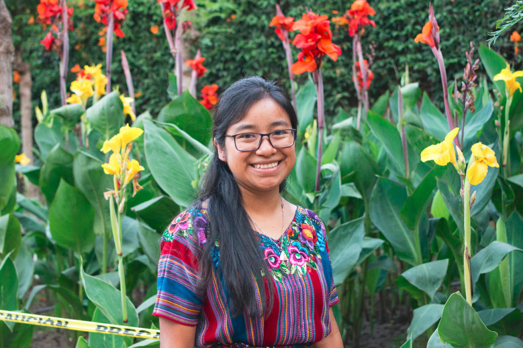 Photograph of a smiling young person in a Mayan huipil in front of bright green, red, and yellow flowers. 