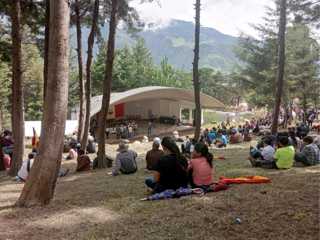 In an open field, in the middle of trees, people sitting on the ground looking towards a white-roofed amphitheater.