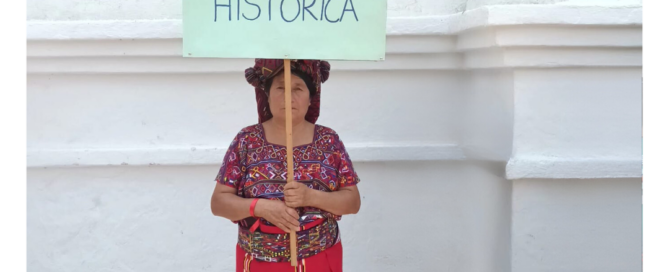 A woman with traditional dress, holds a banner with a spanish quote: The Ixil People have historical memory.
