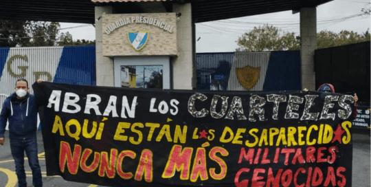 [ESP] Una imagen de una persona socializada hombre, sosteniendo una pancarta de fondo negro con letras blkancas, amarillas y rojas que dice: Abran los cuarteles, aquí estan los archivos. Nunca más militares genocidas. Al fondo la entrada de un cuartel militar [ENG] An image of a male socialized person, holding a black background banner with white, yellow and red letters that reads: Open the barracks, here are the archives. Never again genocidal military. In the background the entrance of a military barracks