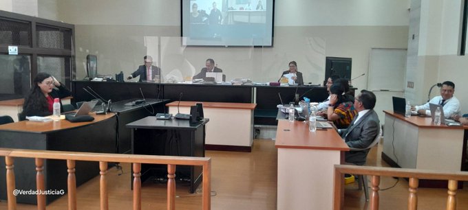 En la foto se observa un tribunal, en los laterales hay mesas donde hay sentadas varias personas hombres y mujeres, al fondo detras de una pantalla de vidrio hay dos hombres y una mujer y detras de ellos una pantalla donde se observan dos personas /// The photo shows a court, on the sides there are tables where several men and women are seated, in the background behind a glass screen there are two men and a woman and behind them a screen where two people can be seen.