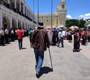 A photograph of Rubén Herrera from behind. He is a tall man walking with dignity and a cane towards a crowd of people. On the right are people holding signs wearing traditional Mayan clothing, on the left a line of police in uniform.
