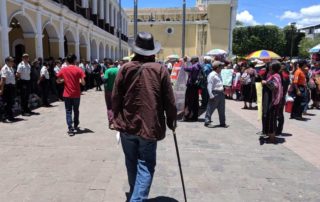 A photograph of Rubén Herrera from behind. He is a tall man walking with dignity and a cane towards a crowd of people. On the right are people holding signs wearing traditional Mayan clothing, on the left a line of police in uniform.