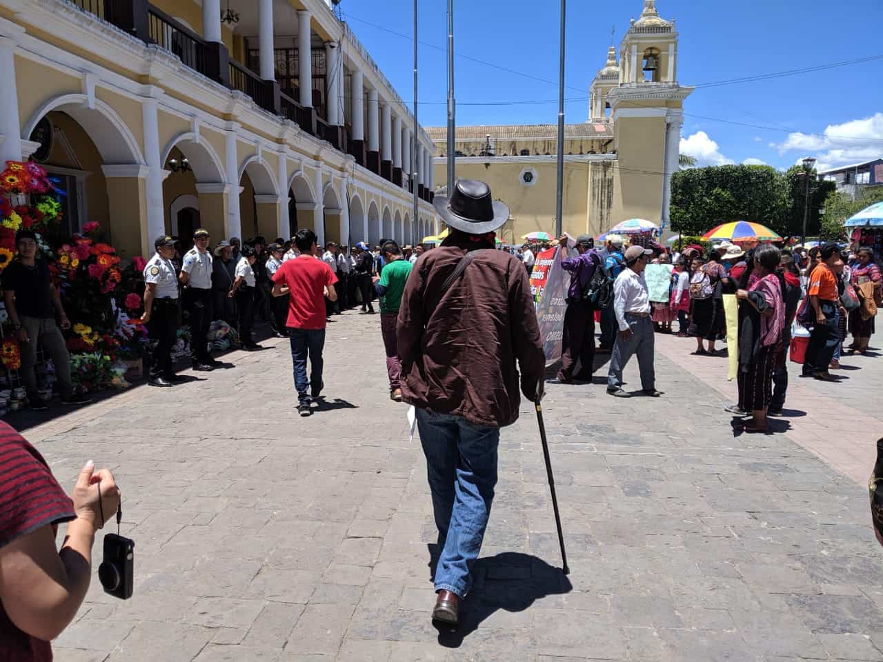 A photograph of Rubén Herrera from behind. He is a tall man walking with dignity and a cane towards a crowd of people. On the right are people holding signs wearing traditional Mayan clothing, on the left a line of police in uniform. 
