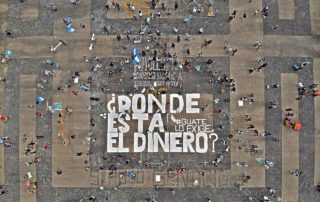 Aerial photo of Plaza de la Constitución in Guatemala City. There are people protesting while social distancing and a couple of Guatemalan flags can be seen. In the center of the picture it is written in white paint “Dónde está el dinero? Guate lo exige”, Where is the Money? Guate Demands it”. The remains of other protests can also be seen, as under as below the main white letters, you can read in black Si hubo genocidio, yes, there was a genocide and the number 56 in yellow, reffering to the murders of the girls in the Hogar Seguro Case.
