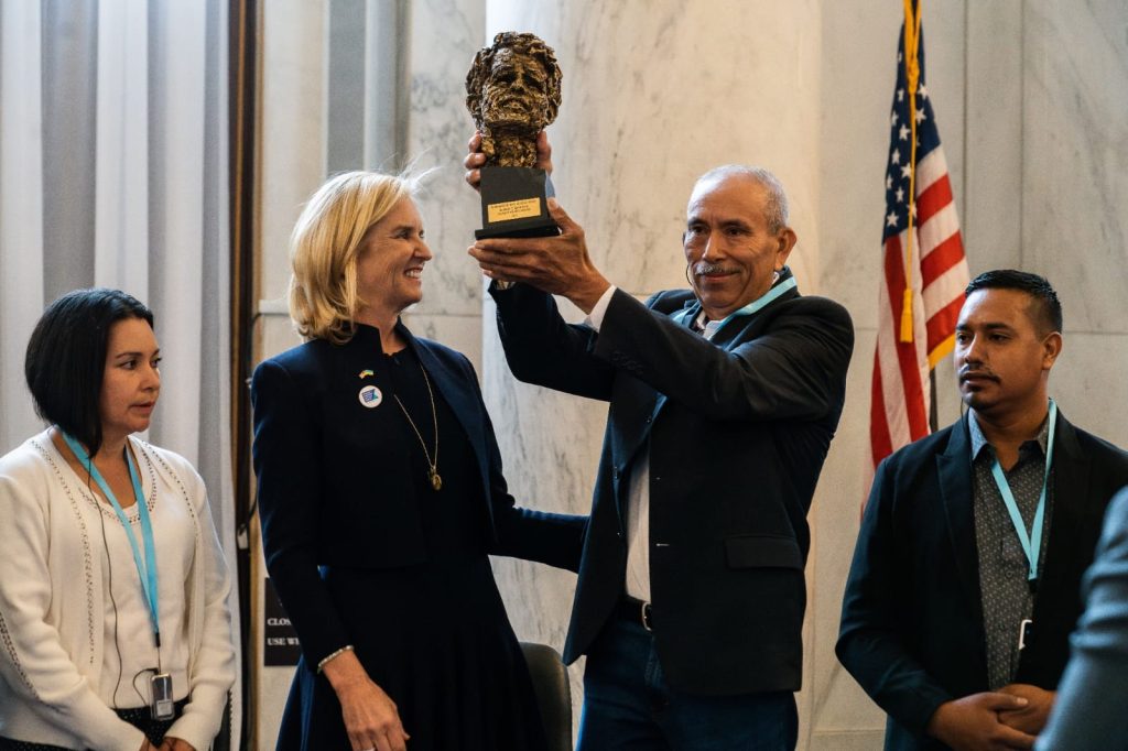 In the photograph there are four people standing, from left to right, there is a woman of medium height with a white sweater, she has short black hair down to her shoulders, next to her is a tall, blonde woman with a navy blue dress, next to her a tall, gray-haired man with a mustache and short hair wearing a black jacket, white shirt and jeans is carrying the trophy which is a metal sculpture in the shape of a face, and next to him a man with a black jacket and gray shirt.