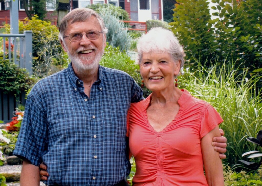 Two people looking straight at the camera, one person socialized as a man with blue checkered shirt, white beard, gray hair and glasses. And the other person socialized as a woman, with pink blouse and white hair. Both hugging and laughing