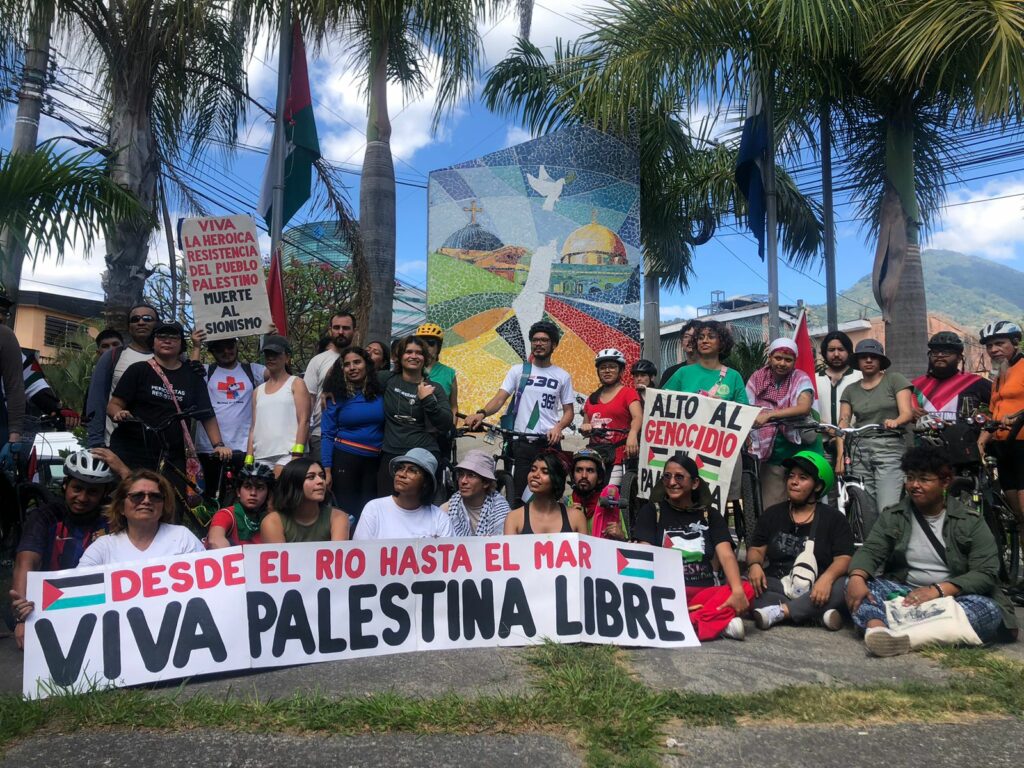 Un grupo de personas reunidos en una plaza. En el fondo un mosaico con iglesias y banderas de El Salvador y Palestina que se enlazan y una paloma blanca. El grupo de personas sostiene una pancarta que dice: “Desde el río, hasta el mar, viva Palestina Libre” 