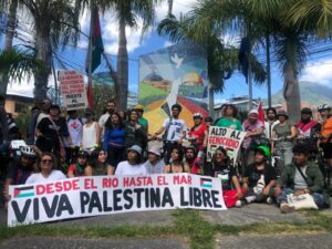 A group of people gathered in a square. In the background is a mosaic with churches and flags of El Salvador and Palestine that are linked and a white bird. The group of people hold a banner that says: "From the river to the sea, long live Free Palestine".