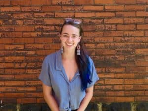 Photo of Zia Kandler smiling in front of a brick wall. She is a white femme with long brown hair with blue tips.