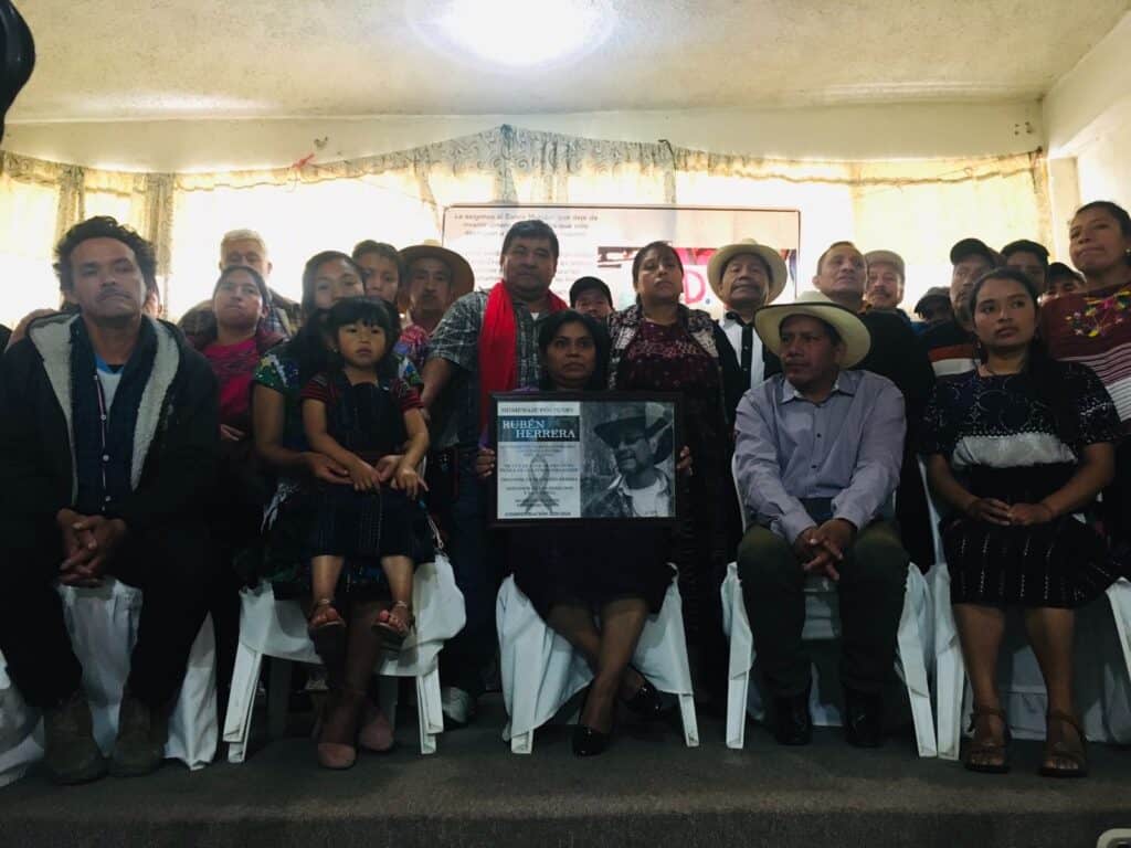 he image shows a group of 20 people huddled together, many wearing their traditional mayan clothing, and at the front a woman holding a black and white photograph of the late organizer Ruben Herrera.