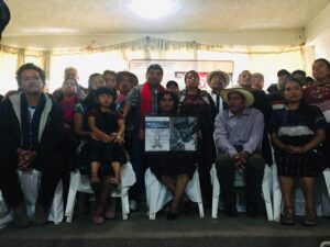 he image shows a group of 20 people huddled together, many wearing their traditional mayan clothing, and at the front a woman holding a black and white photograph of the late organizer Ruben Herrera.