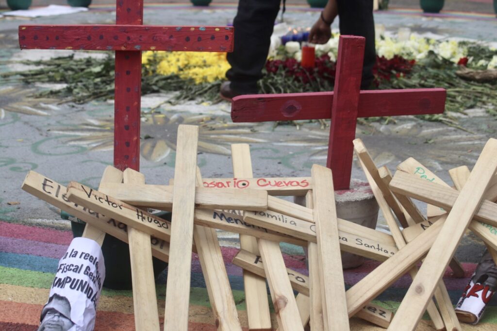 Several wooden croses with names on them are on the floor. Standing there are a few red crosses, behind the crosses you can see a man taking care of a flower altar