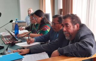 traduce, en la imagen se observa una sala de audiencias, en primer plano cuatro abogados, tres hombres y una mujer. los hombres usan traje y corbata gris, la mujer tiene vestimenta tradicional. En el fondo se observa otra persona de camisa azul clara. En el escritorio se observan documentos y computadoras In the image, a courtroom is observed. In the foreground, there are four lawyers: three men and one woman. The men are wearing gray suits and ties, while the woman is dressed in traditional attire. In the background, another person can be seen wearing a light blue shirt. On the desk, there are documents and computers visible.