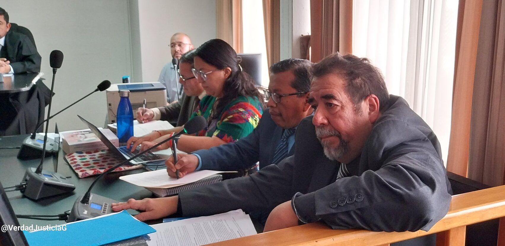 traduce, en la imagen se observa una sala de audiencias, en primer plano cuatro abogados, tres hombres y una mujer. los hombres usan traje y corbata gris, la mujer tiene vestimenta tradicional. En el fondo se observa otra persona de camisa azul clara. En el escritorio se observan documentos y computadoras In the image, a courtroom is observed. In the foreground, there are four lawyers: three men and one woman. The men are wearing gray suits and ties, while the woman is dressed in traditional attire. In the background, another person can be seen wearing a light blue shirt. On the desk, there are documents and computers visible.