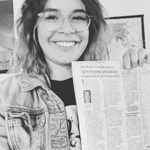 Photograph of a smiling femme with glasses holding a newspaper. The headline reads "Asylum Cooperative Agreements should be suspended permanently"