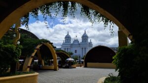 Arriba hay una arca con plantas colgando, enmarcando un cielo azul con nubes, un parque central con otros arcos verdes y una iglesia blanca con revestimiento azul y dos campanarios con cúpula.