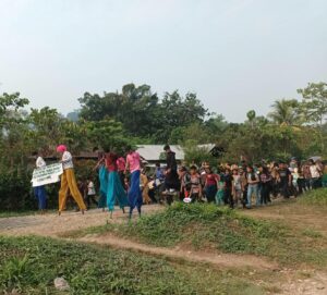 About 50 youth walk down a road lined with trees. Some are on stilts and some wear costumes. They carry signs about contamination of the earth and water. Unos 50 jóvenes caminan por una carretera bordeada de árboles. Algunos van sobre zancos y otros llevan disfraces. Llevan carteles sobre la contaminación de la tierra y el agua.