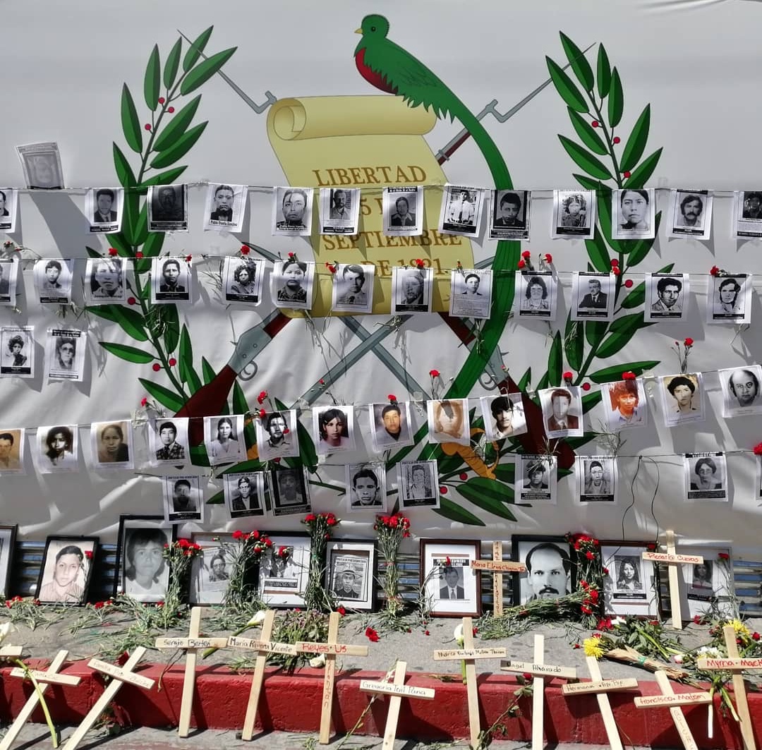 Covering a giant poster with guatemalan national symbols, organizations put in the front pictures of the dissapeared and murder during the Internal Armed Conflict along with red carnations and crosses