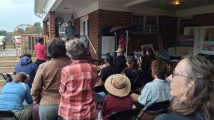 The scene depicts an outdoor community event. A group of people sit and stand facing a speaker on the porch of a brick building. The attendees are dressed casually, some wearing jackets and hats.