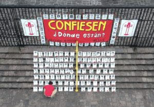Outside a fence there is a sign that reads “Confess: Where are they?” On the stairs, a person with a red t-shirt is laying the black and white pictures of the 183 disappeared who are registered in the Military Diary