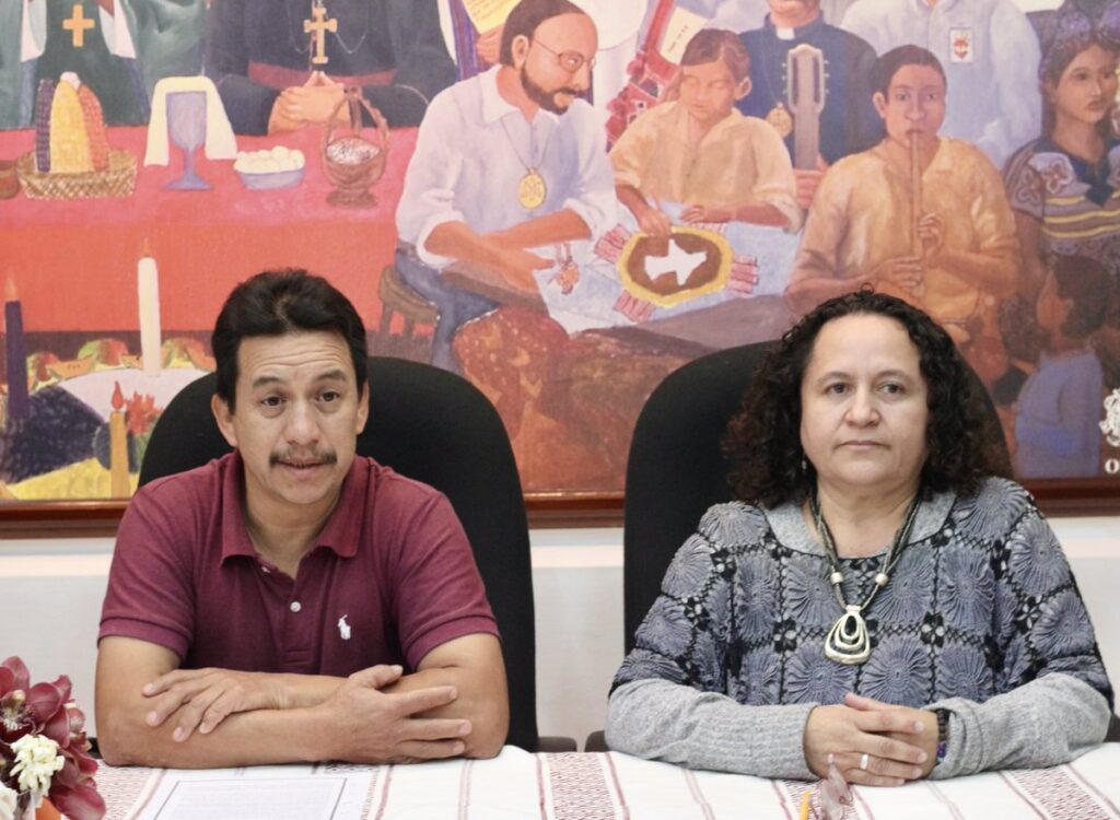 In the background a mural depicting the work of church members and martyrs during the internal armed conflict shows men in civilian clothes or priests doing various activities, playing music, making posters or preparing materials for mass. In the foreground on the left side you can see a flower arrangement, then there is a brown middle-aged man with his arms crossed and a red t-shirt. On the right side there is a middle-aged woman with curly hair, dressed in gray and with her hands intertwined./////Al fondo un mural que representa el trabajo de miembros y martires de la iglesia durante el conflcito armado interno se puede ver a hombrs vestidos de civil o curas hciendo diversas actividades, tocando musica, haciendo carteles o preparando matertiales para la misa. En primer plano de lado izquierdo se alcanza a ver un arreglo floral, despues se encuentra un hombre moreno de edad mediana con los brazos cruzados y una playera roja. del lado derecho se encuentra una mujer de edad mediana con cabbello rizado y esta vestida de gris y tiene sus manos entrelazadas