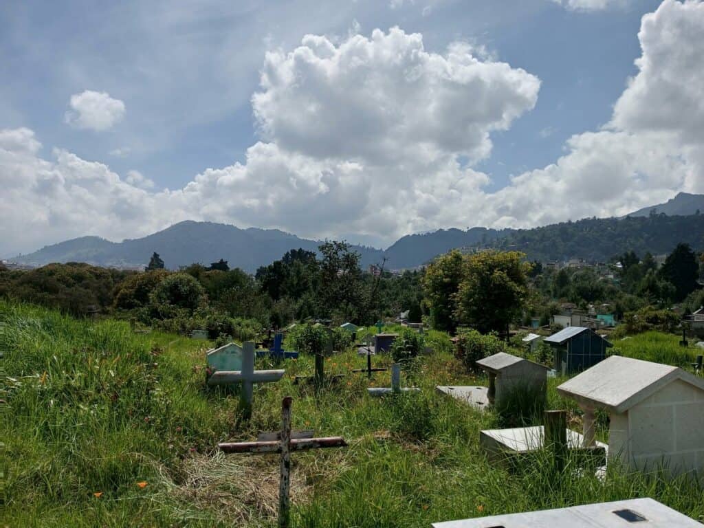 view of a cemetery with several crosses and graves in a field. Mountains and a cloud sky are in the background