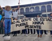 Manifestantes sostienen una manta blanca con texto negro que dice "No al golpe de estado, Pueblo Xinka presente" frente a un edificio colonial con arcos y el letrero "Corte de Constitucionalidad". Un hombre con sombrero de ala ancha y camisa a cuadros azules levanta los brazos sosteniendo un bastón ceremonial mientras está parado sobre una esfera de concreto.
