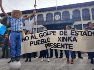 Manifestantes sostienen una manta blanca con texto negro que dice "No al golpe de estado, Pueblo Xinka presente" frente a un edificio colonial con arcos y el letrero "Corte de Constitucionalidad". Un hombre con sombrero de ala ancha y camisa a cuadros azules levanta los brazos sosteniendo un bastón ceremonial mientras está parado sobre una esfera de concreto.