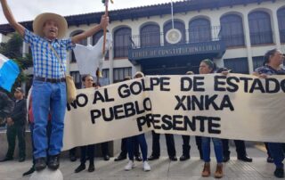Manifestantes sostienen una manta blanca con texto negro que dice "No al golpe de estado, Pueblo Xinka presente" frente a un edificio colonial con arcos y el letrero "Corte de Constitucionalidad". Un hombre con sombrero de ala ancha y camisa a cuadros azules levanta los brazos sosteniendo un bastón ceremonial mientras está parado sobre una esfera de concreto.