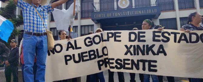 Manifestantes sostienen una manta blanca con texto negro que dice "No al golpe de estado, Pueblo Xinka presente" frente a un edificio colonial con arcos y el letrero "Corte de Constitucionalidad". Un hombre con sombrero de ala ancha y camisa a cuadros azules levanta los brazos sosteniendo un bastón ceremonial mientras está parado sobre una esfera de concreto.