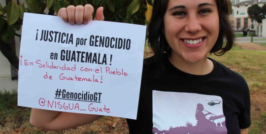 A photograph of a NISGUA organizer holding a sign that reads "Justicia por genocidio en Guatemala!"