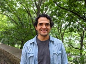 A photograph of Miguel, a smiling masc person with black hair, grey shirt, and a loose jean jacket.