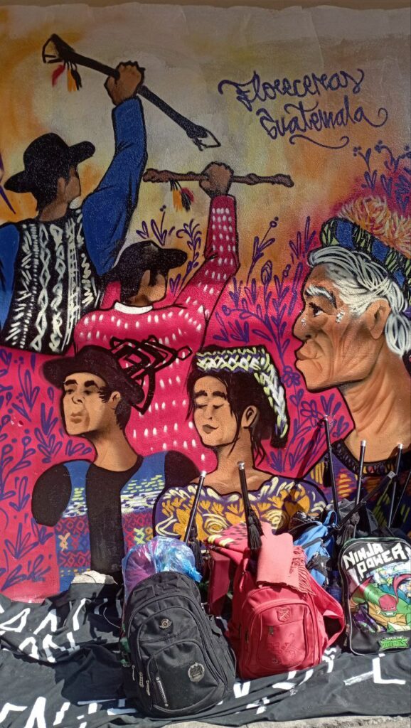 En la imagen están pintados con graffiti cinco personas de los pueblos originarios, una abuela, una mujer y tres hombres, dos de ellos de lado y sosteniendo cada uno de ellos su vara de mando y la frase “Florecerás Guatemala”, recargadas en el muro están cinco mochilas con varias varas de mando guardadas.