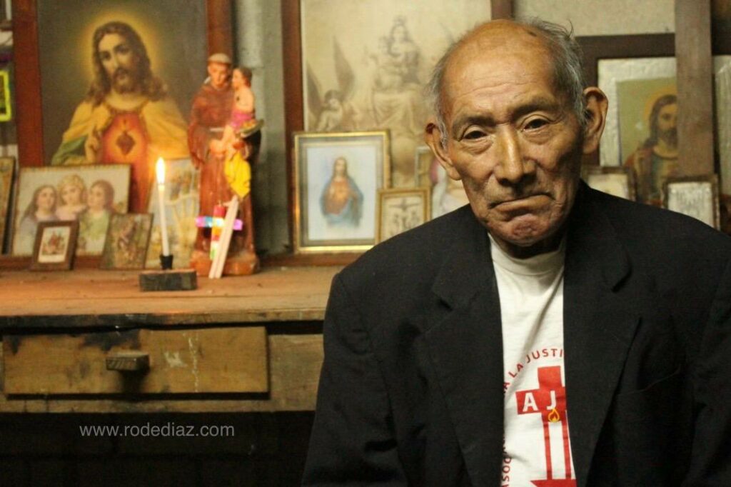 A person socialized as a man. Elder. White shirt with AJR red logo and a black jacket. In the background pictures, prints and lumps of catholic figures.