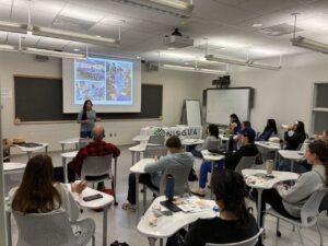 A bright classroom is full of some three dozen people sitting on classroom desks face forward toward a presenter wearing pink clothing and speaking to the large audience. The classroom walls are covered witth art and TV screen projecting a presentation.