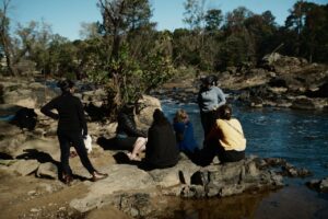 Un pequeño grupo de cinco personas está sentado y de pie sobre unas grandes rocas junto a un río. Una persona habla mientras las demás escuchan. Algunas llevan auriculares para la interpretación lingüística.
