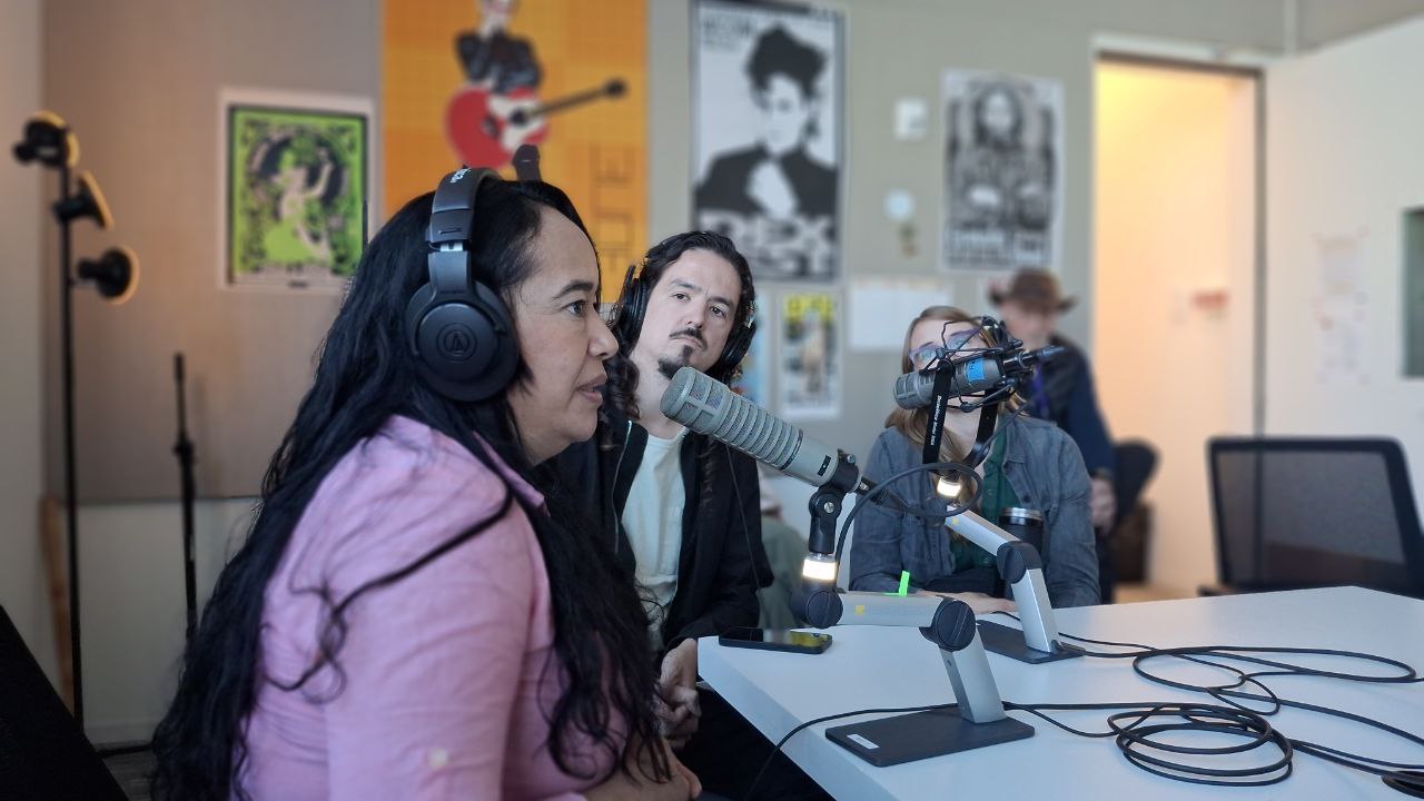 Una mujer con una camiseta rosa lleva unos audífonos y habla por un micrófono profesional en lo que parece ser un estudio de radio. Tres personas observan a la oradora desde el fondo. Las paredes están cubiertas con imágenes borrosas de personas con instrumentos.