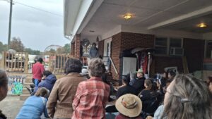 La escena muestra un evento comunitario al aire libre. Un grupo de personas está sentado y de pie frente a un ponente en un edificio de ladrillo. Los participantes visten ropa informal, algunos con chumpas y gorras.