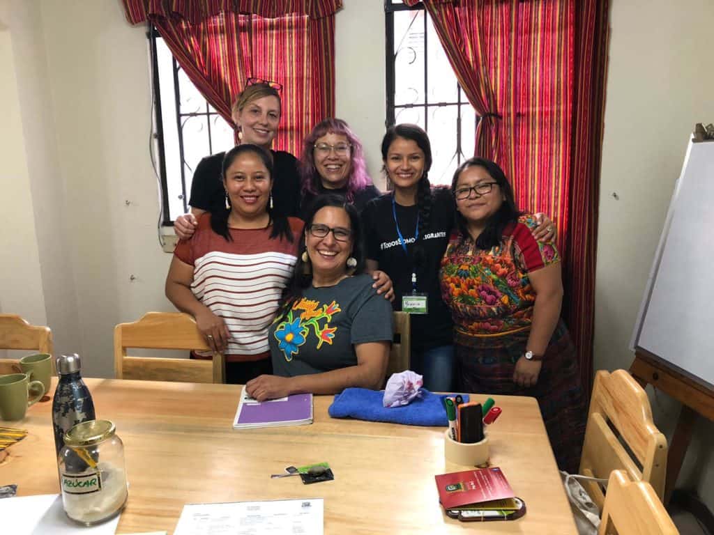 Six women smile in a tight group in front of open windows. 