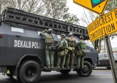 ESP: Sobre una calle un tanque policial con cuatro personas agarradas a un costado del vehículo, con vestimenta verde militar, cascos, chalecos antibalas y armas. ENG: On a street, a police tank with four people clinging to the side of the vehicle, wearing green military clothing, helmets, shawls