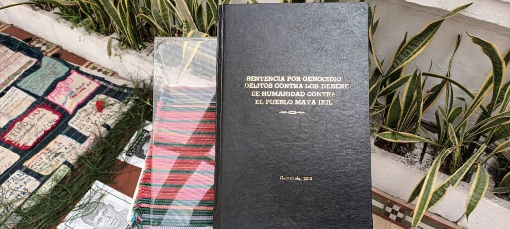 ESP: En el fondo del lado izquierdo se mira el borde de una manta color azul sobre ella retazos de manta beige con nombres de víctimas asesinadas y desaparecidas durante el conflicto armado. En el lado derecho sobre la manta, en un podio transparente un libro con cobertura negra con letras doradas que dice: “Sentencia por Genocidio. Delitos contra los deberes de humanidad contra el Pueblo Maya Ixil” ENG: In the background on the left side you can see the edge of a blue blanket with patches of beige blanket with names of victims killed and disappeared during the armed conflict. On the right side of the blanket, on a transparent podium, a book with a black cover and golden letters that reads: "Sentence for Genocide. Crimes against the duties of humanity against the Maya Ixil People".