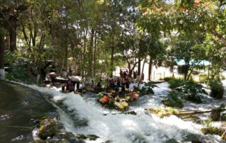 The photograph shows the beginning of the San Juan River. The river is flowing from left to right in the image’s foreground and is shaded by small trees. In the middle of the river there are many bundles of flowers used as offerings in Mayan ceremonies. In the background there is a group of Indigenous people praying.