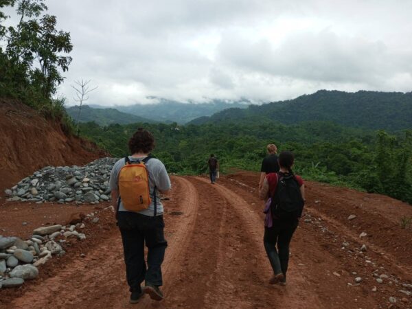 Group of people walking along a red dirt road under construction through a mountainous green landscape. Piles of stones are visible on the sides of the road, with forested hills covered in lush tropical vegetation and a cloudy sky. The walkers are carrying backpacks and the scene suggests a rural developing area with misty mountains in the background.